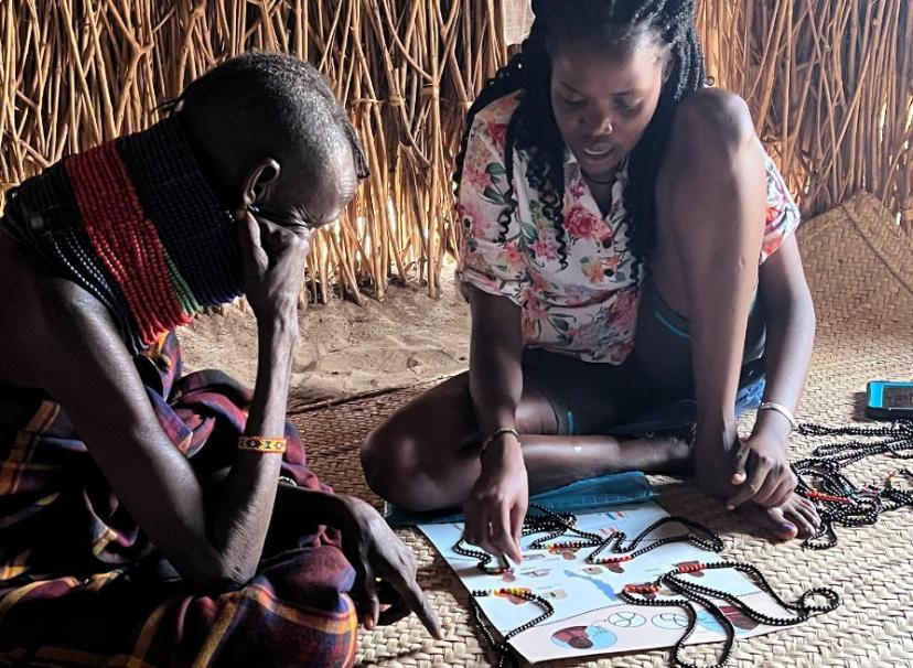 Two women are sitting down on the floor. The woman on the right points to a diagram on a poster about DNA. There are strings of black and colored beads scattered across the poster and floor.